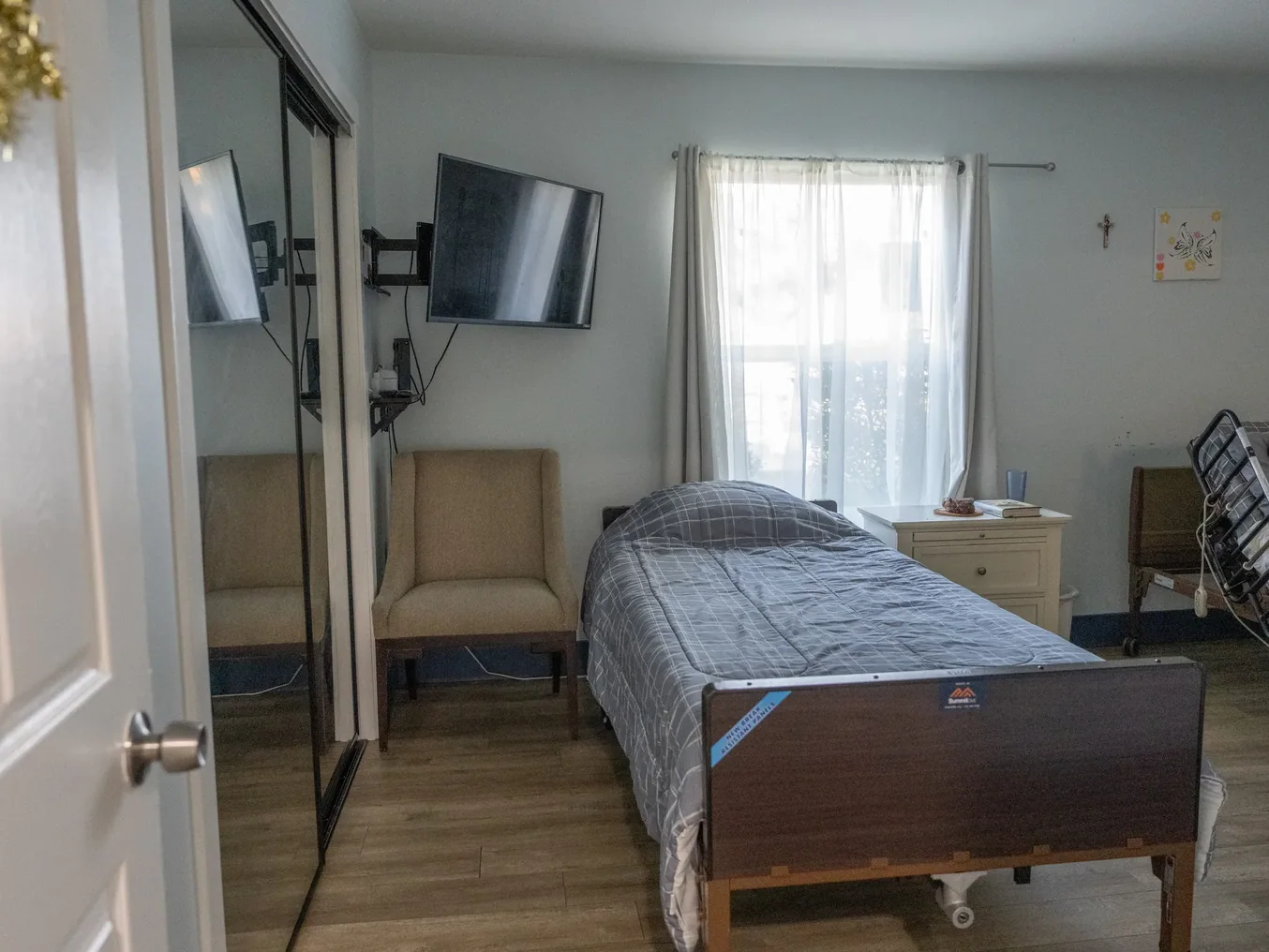 Residential care bedroom featuring hospital bed, comfortable seating, entertainment system, and accessible furnishings with natural lighting